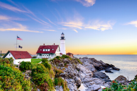A scenic lighthouse sits on a rocky shore, complemented by a white and red-roofed building nearby, with flags waving in the gentle breeze and a calm sea stretching to the horizon under a colorful sunset sky.
