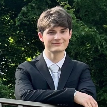 A young man in a black suit with a white shirt and silver tie, smiling and leaning on a railing, surrounded by green foliage.