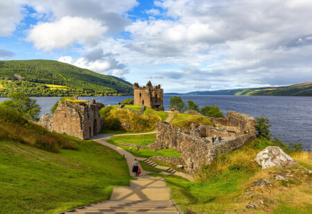 A scenic view of the ruins of a castle situated on a hillside by a large lake, surrounded by green hills under a partly cloudy sky, with visitors walking along a winding path.