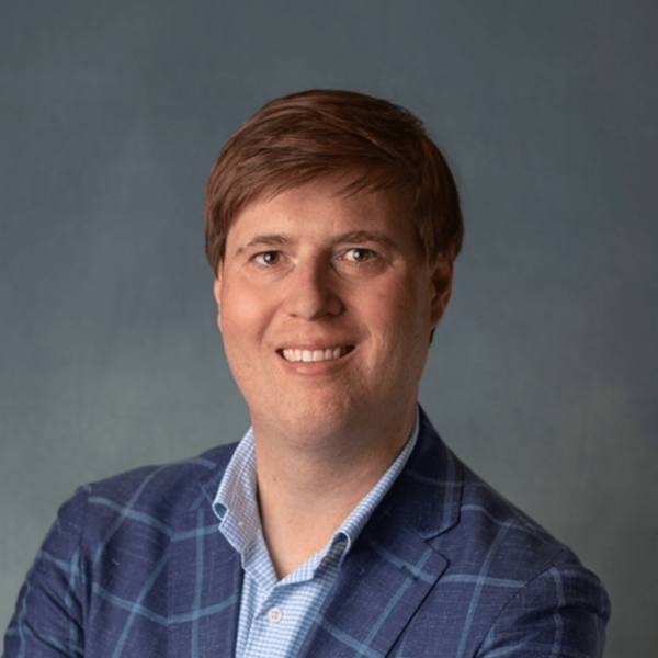 A smiling man with medium-length brown hair wearing a blue plaid blazer over a light blue shirt, standing against a soft gray background.