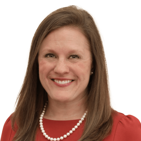 A woman with long brown hair is smiling while wearing a red dress and a pearl necklace against a plain white background.