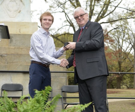 A young man in a white shirt shakes hands with a man in a dark suit, both smiling, during a formal event outdoors. They are positioned in front of a stone monument, with several folding chairs and greenery in the foreground.