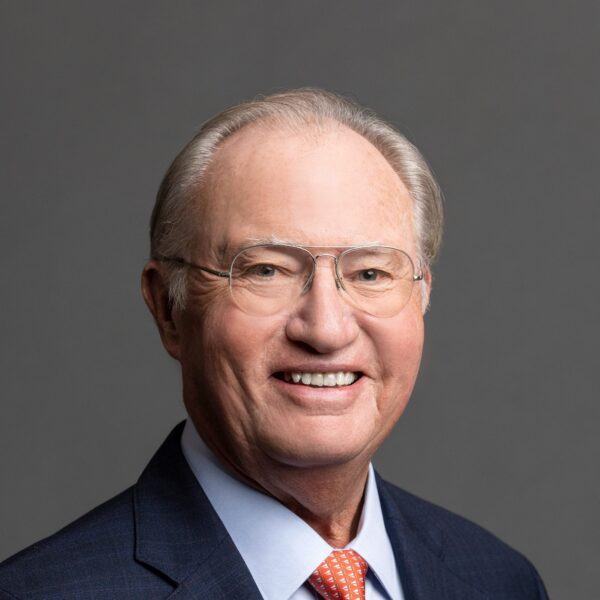 A smiling older man with glasses and light-colored hair, wearing a dark blue suit and an orange tie, poses against a grey background.