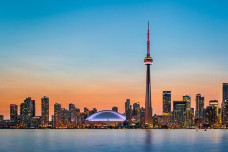 The Toronto skyline at sunset, featuring the iconic CN Tower illuminated in red and white, with the Rogers Centre stadium in the foreground and numerous skyscrapers in the background against a gradient sky from orange to blue.