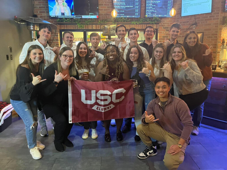 A group of young adults poses together in a casual bar setting, holding a USC Alumni flag. They are making hand gestures with smiles, with a mix of drinks visible on the table. The background features a bar area and illuminated screens.