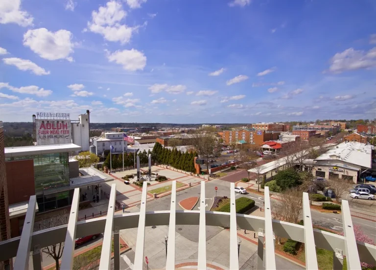 A panoramic view of a town square with modern buildings and a historic mill visible in the foreground, accompanied by a clear blue sky filled with white clouds. The view includes a landscaped area with trees, a flagpole, and several roads lined with parked cars.