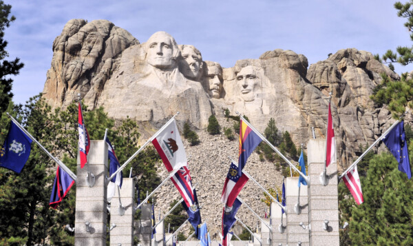 Mount Rushmore featuring the carved faces of four U.S. presidents, surrounded by flagpoles displaying various state flags and trees in the foreground.