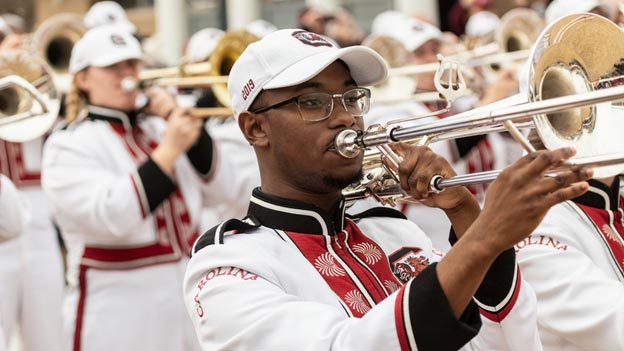 A musician in a marching band plays a trombone, wearing glasses and a white hat with 2019 printed on it, dressed in a white and black uniform with red accents. Other band members are visible in the background, also playing brass instruments.