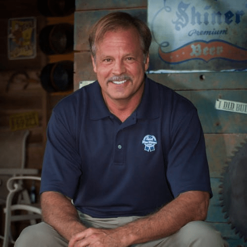 A smiling middle-aged man with short brown hair, wearing a dark blue polo shirt with a logo, sitting against a rustic wooden backdrop decorated with signs and beer-related items.