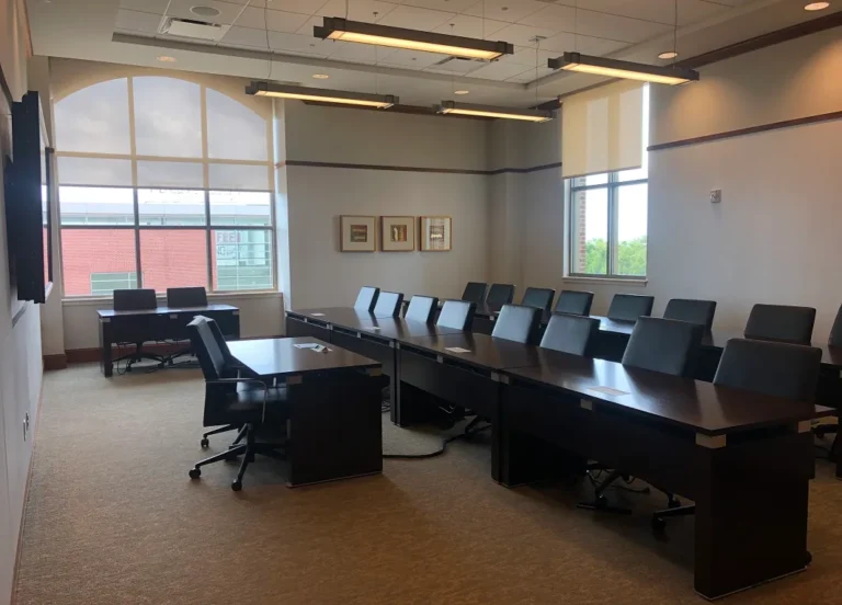A modern conference room featuring a long table with black chairs on either side, a smaller table at the front, a large screen on the wall, and large windows with shades, providing natural light.