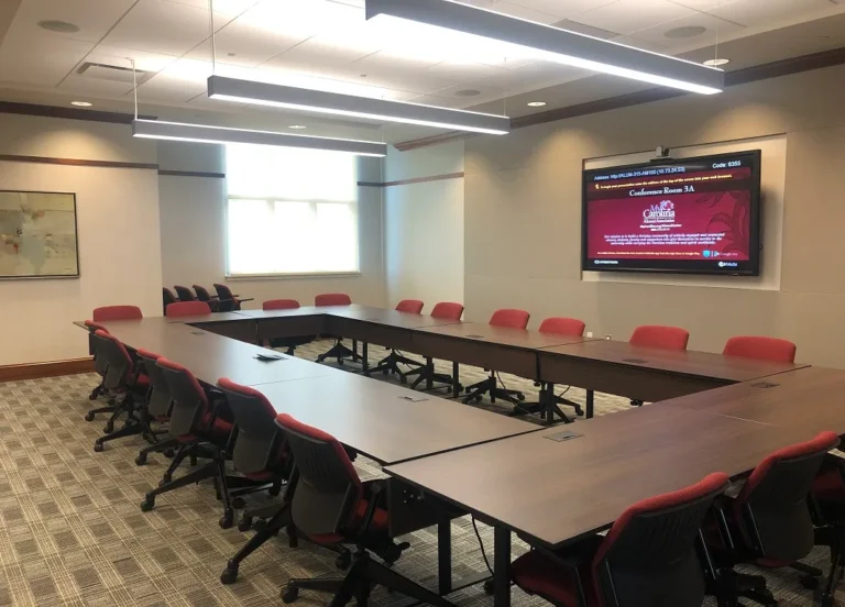 A conference room featuring a large rectangular table surrounded by red ergonomic chairs, with a large screen displaying information on the wall. The room is well-lit with modern lighting and windows allowing natural light to enter.
