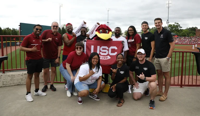 A group of alumni from the University of South Carolina, posing together with the school's mascot, holding a USC Alumni banner, at a baseball game.