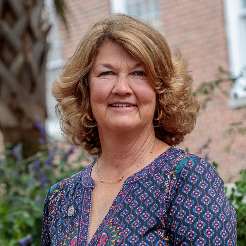A woman with medium-length curly hair smiles warmly, wearing a colorful patterned blouse, set against a background of greenery and brick architecture.
