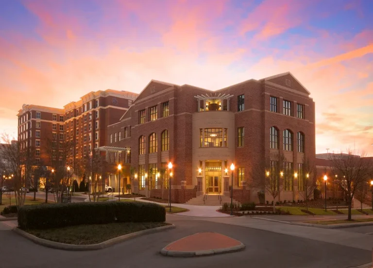 A modern brick building with large windows and a grand entrance, surrounded by landscaped greenery and street lamps, set against a colorful sunset sky. A larger building is visible in the background.