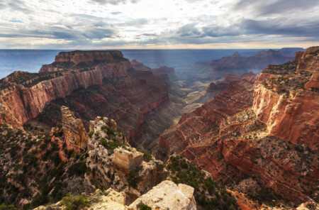 A panoramic view of the Grand Canyon showing dramatic red and orange rock formations, layered cliffs, and a valley below, with a cloudy sky above casting soft light on the landscape.