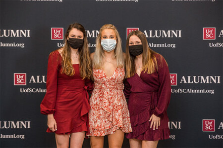 Three women posing together in front of a backdrop with 'UofSC ALUMNI' and website text, all wearing masks. The woman on the left wears a red dress with long sleeves, the central woman wears a floral dress, and the woman on the right wears a deep red dress, all smiling.