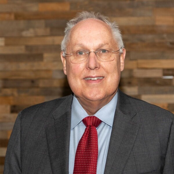 A smiling older man with glasses wearing a gray suit and red tie, standing in front of a textured wooden wall.