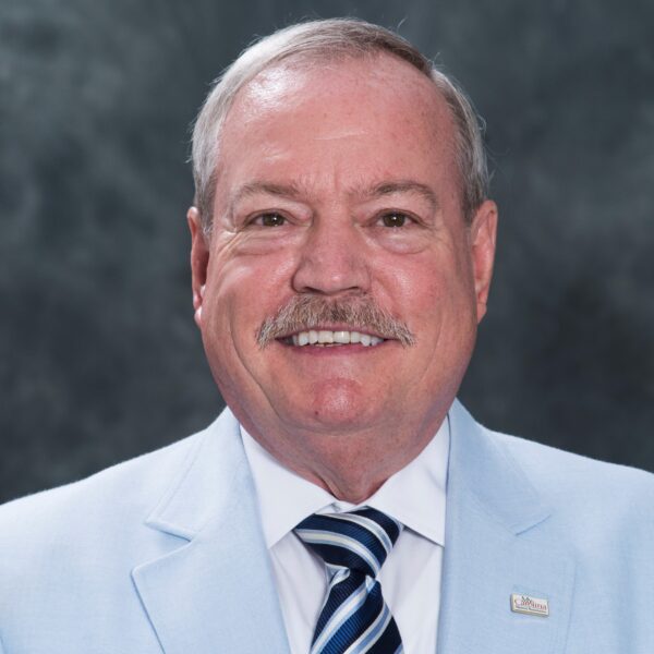 A man with a mustache is smiling at the camera, wearing a light blue suit with a white shirt and striped tie. He has short hair and is standing against a dark textured background.