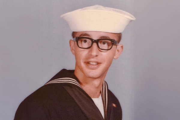 A young man in a navy uniform with a white sailor hat and black-rimmed glasses, smiling and looking off to the side, against a soft blue background.