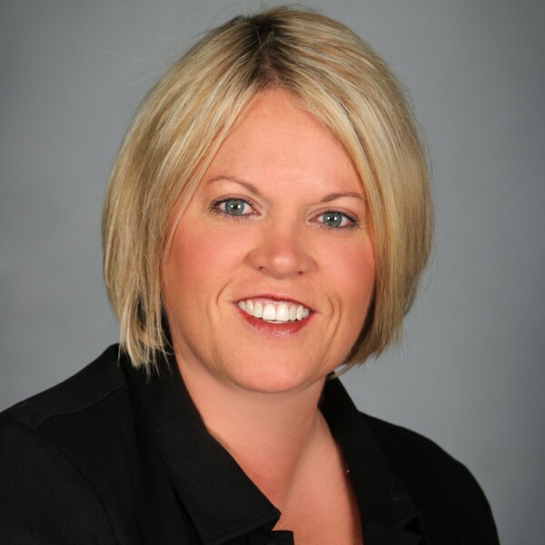 A smiling woman with short blonde hair wearing a black jacket, against a neutral gray background.