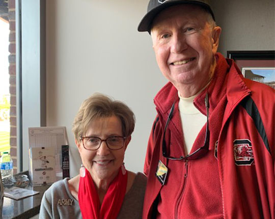 Two smiling older adults pose for a photo indoors; the woman, with short brown hair and glasses, wears a gray sweater with 'ARMY' embroidered and a red scarf, while the man, with gray hair and a light-colored shirt, wears a red jacket with a logo and a black cap.