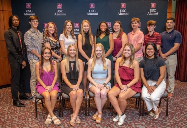 A group photo of 15 young adults standing and sitting in front of a USC Alumni backdrop, featuring a diverse mix of individuals, some wearing formal attire and others in casual clothing, arranged in two rows with smiles and posed for the camera.