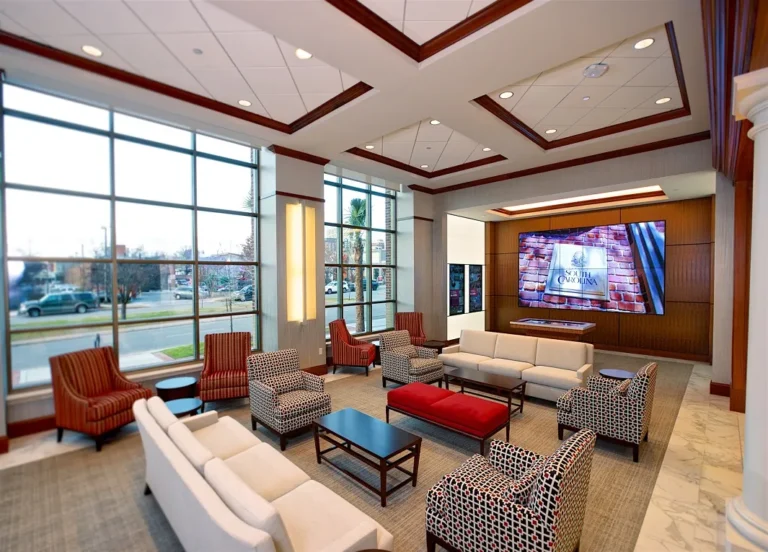A spacious lobby with large windows, featuring multiple seating arrangements including white sofas, red accent chairs, and patterned armchairs. A large screen displays an image of South Carolina on a wooden wall, while a coffee table sits centrally on a textured rug.