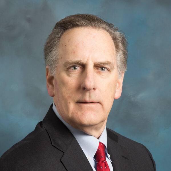 A serious-looking man with short gray hair and light skin, wearing a dark suit with a light blue shirt and a red tie, is facing the camera against a softly blurred blue background.