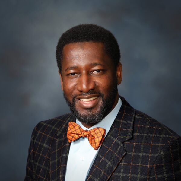 A smiling man with a beard wearing a dark plaid suit jacket and an orange bow tie over a light blue shirt, set against a softly blurred blue background.