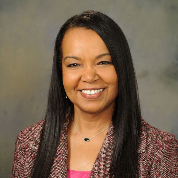 A woman with long, straight black hair smiles at the camera, wearing a pink top and a textured pink blazer. She is adorned with a silver necklace featuring a black stone, and her expression is warm and approachable against a soft, muted background.