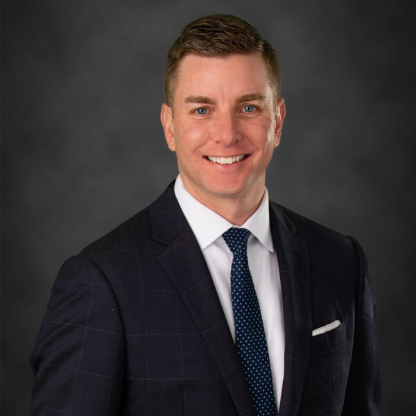A smiling man in a dark suit with a white shirt and a blue polka dot tie, posing against a gradient gray background.