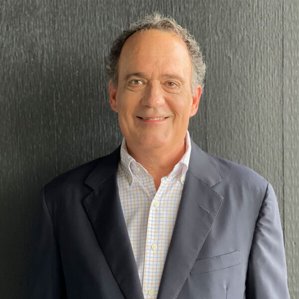 A man with curly hair wearing a navy blazer and a light checkered shirt, smiling warmly, against a textured dark background.