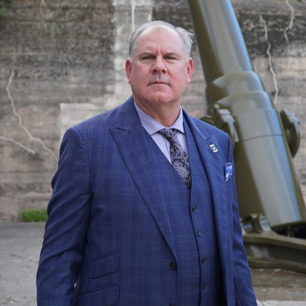 A serious-looking man in a dark blue plaid suit stands confidently, with a purple dress shirt and patterned tie. In the background, there is a large green artillery piece against a textured stone wall.