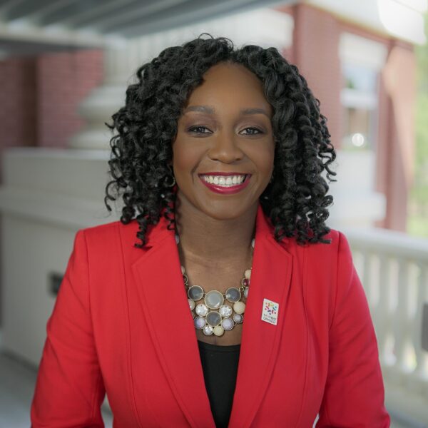 A smiling woman with curly black hair, wearing a red blazer and a statement necklace, stands in front of a light background, likely outdoors or in a brightly lit building.