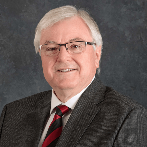 A smiling older man with gray hair and glasses, wearing a dark blazer over a white shirt with a red and black striped tie, set against a textured gray background.