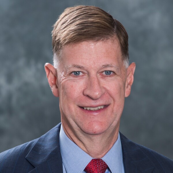 A formal headshot of a man with short, neatly combed hair, wearing a blue suit and a light blue shirt, with a red tie. He is smiling and has blue eyes, against a soft, blurred gray background.