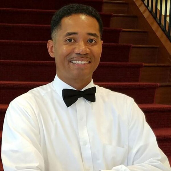 A man smiling while wearing a white dress shirt and a black bow tie, standing on a staircase with red carpeting in the background.