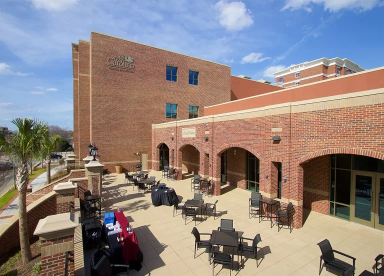 A courtyard area in front of a brick building labeled 'My Carolina Alumni Center,' featuring several outdoor tables and chairs, some covered with black and burgundy tablecloths, and palm trees along the edges. The sky is clear with scattered clouds.