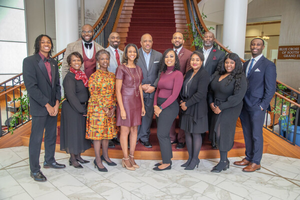 A group portrait of thirteen diverse individuals posing on a staircase with a polished wooden railing, dressed in formal attire. They stand in two rows; the background features a beautifully designed interior, with a tile floor and decorated plants.