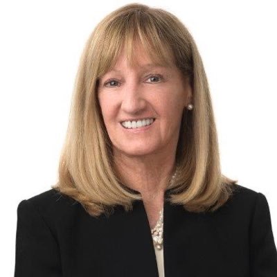 A woman with long, straight blonde hair smiles at the camera, wearing a black blazer and a pearl necklace, against a plain white background.