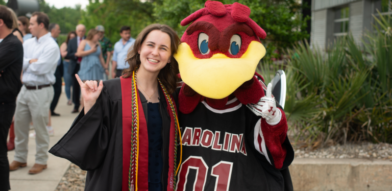 A graduate in a black gown with colorful stoles poses with a red and yellow mascot in a black jersey marked 'CAROLINA 01'. They are outdoors with people milling about in the background.