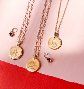A collection of gold necklaces displayed on a pink and red background, featuring two pendants with intricate designs and a small earring with a gemstone.