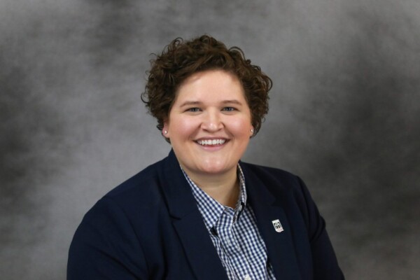 A smiling individual with short, curly hair wearing a dark blazer over a checkered shirt. They are set against a blurred gray background, exuding a friendly demeanor.