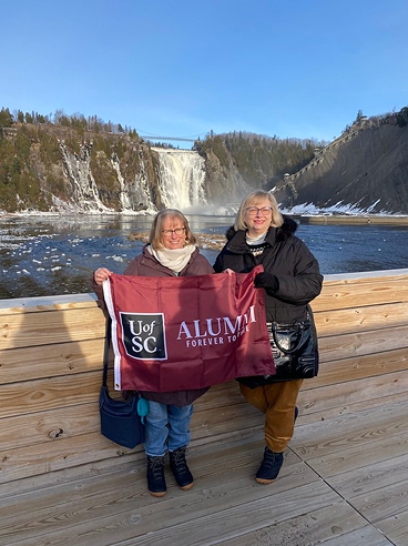 Two women standing on a wooden platform by a frozen waterfall, holding a maroon flag that reads 'UofSC ALUMNI FOREVER TOGETHER', with winter scenery in the background.