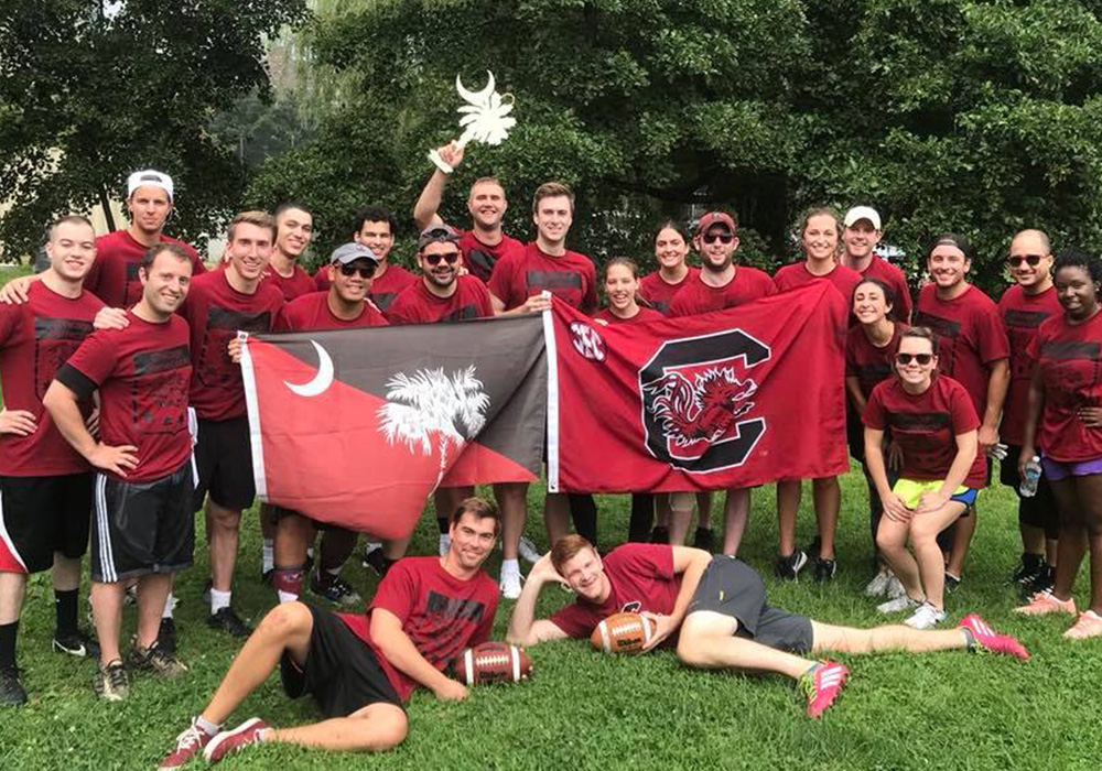 A group of about 25 people wearing red t-shirts posing together outdoors, some holding a flag with a South Carolina design, and one person sitting on the grass holding a football. The scene is set in a lush green park.