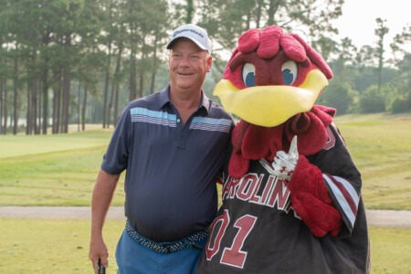 A smiling man stands on a golf course next to a large, costumed mascot that resembles a red gamecock, both posing for a photo. The man is wearing a blue polo shirt and blue shorts, holding a golf club in his left hand, while the mascot is dressed in a jersey with 'CAROLINA' written on it. Trees are visible in the background.