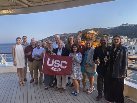 A group of alumni poses on a deck with a view of the sea and hillside in the background, holding a USC Alumni banner. The group, wearing a mix of formal and casual attire, includes men and women of various ages, some holding glasses of beverages.