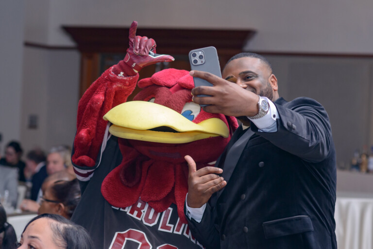 A man in a black suit poses for a selfie with a red bird mascot wearing a jersey, both smiling and making hand gestures, in a banquet setting with blurred people in the background.