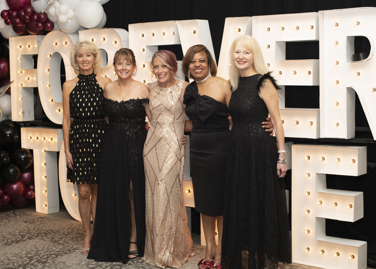 Five women pose together in elegant dresses in front of large illuminated letters spelling 'FOREVER'. The backdrop features decorative balloons in shades of black and burgundy.