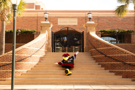 A colorful mascot dressed in a red and black outfit sitting on the steps of the Alumni Center, which has a brick facade and elaborate iron gates with a design featuring a spartan helmet, surrounded by palm trees and festive decorations.